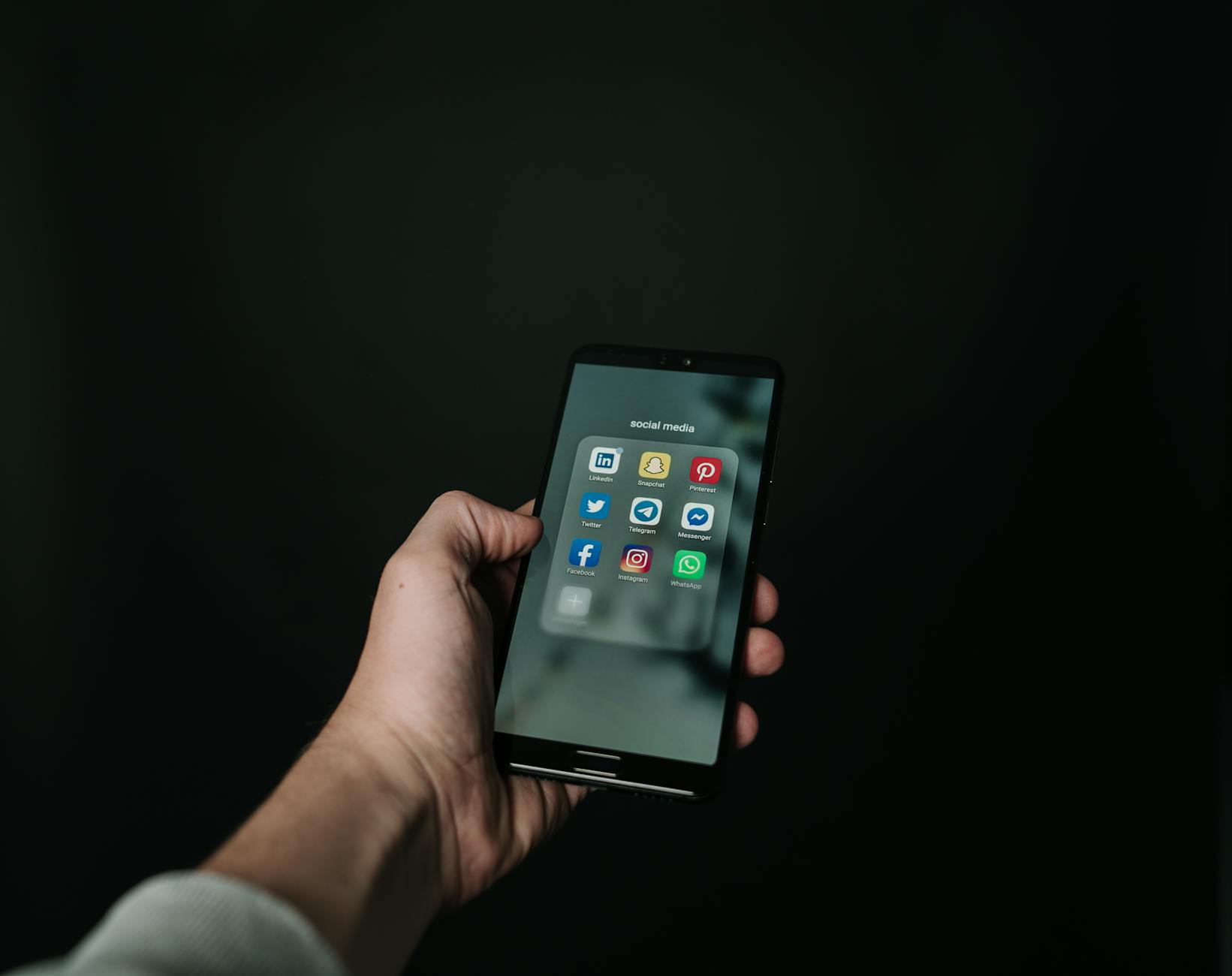 Close-up of a hand holding a smartphone displaying various social media app icons on a dark background